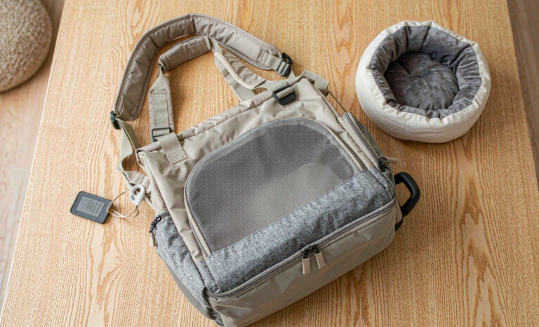 Soft-sided cat travel carrier bag with mesh window next to a plush cat bed on a wooden table